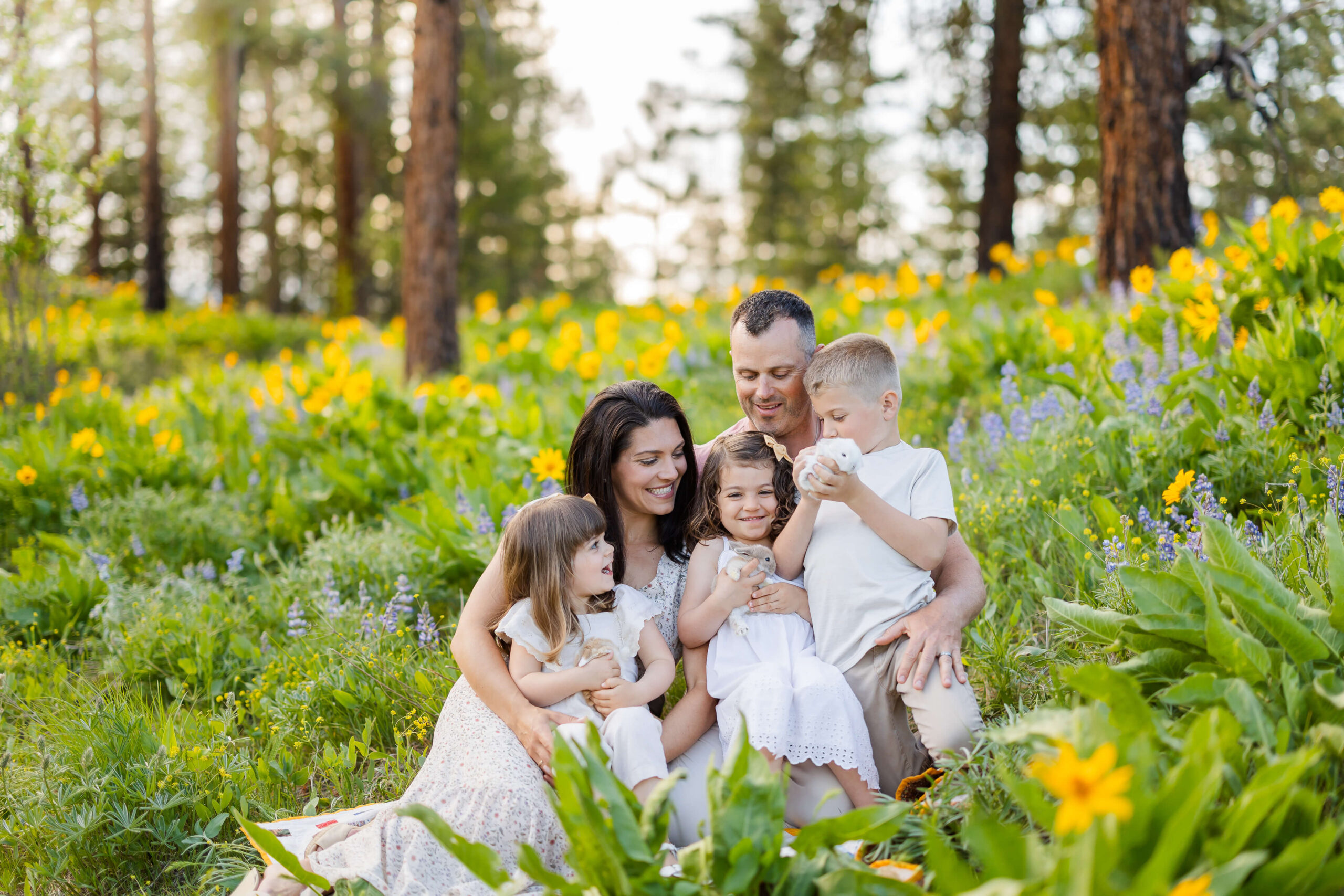 A happy family plays with baby bunnies in mom and dad's lap in a forest filled with yellow and blue wildflowers