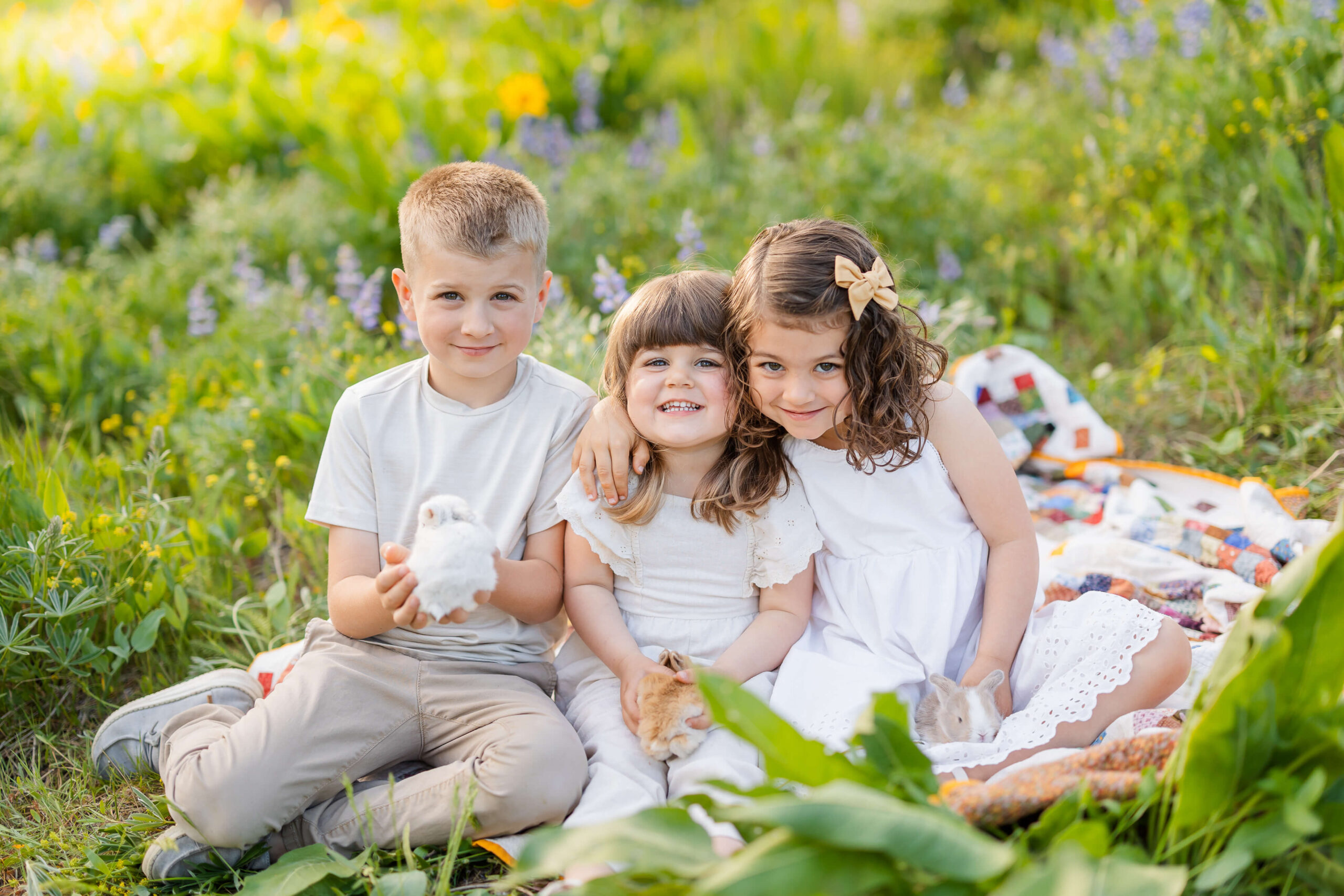 Three young siblings sit in a meadow of wildflowers holding baby bunnies and each other in white outfits after visiting Leavenworth Art Haus