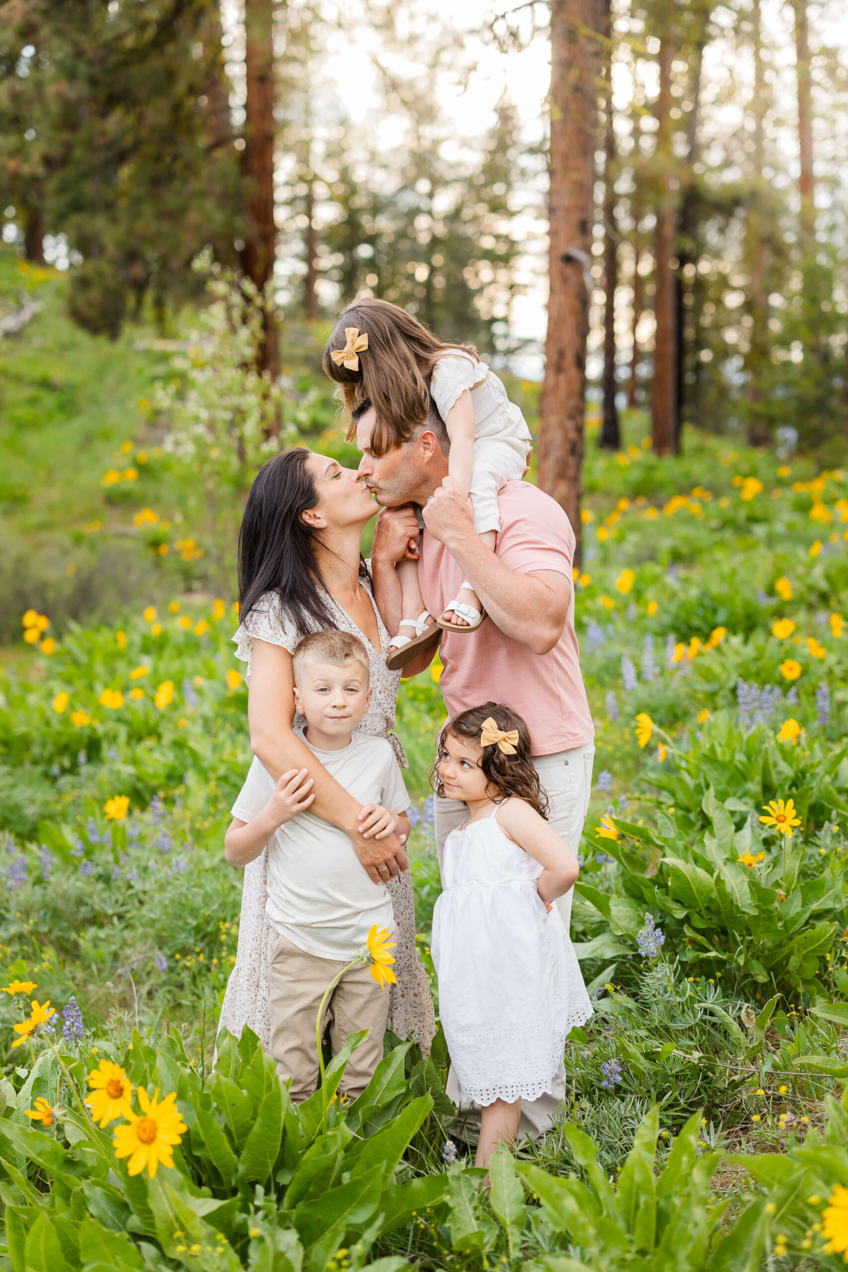 A mom and dad kiss with their three toddlers in a forest filled with wildflowers at sunset after visiting Leavenworth Art Haus