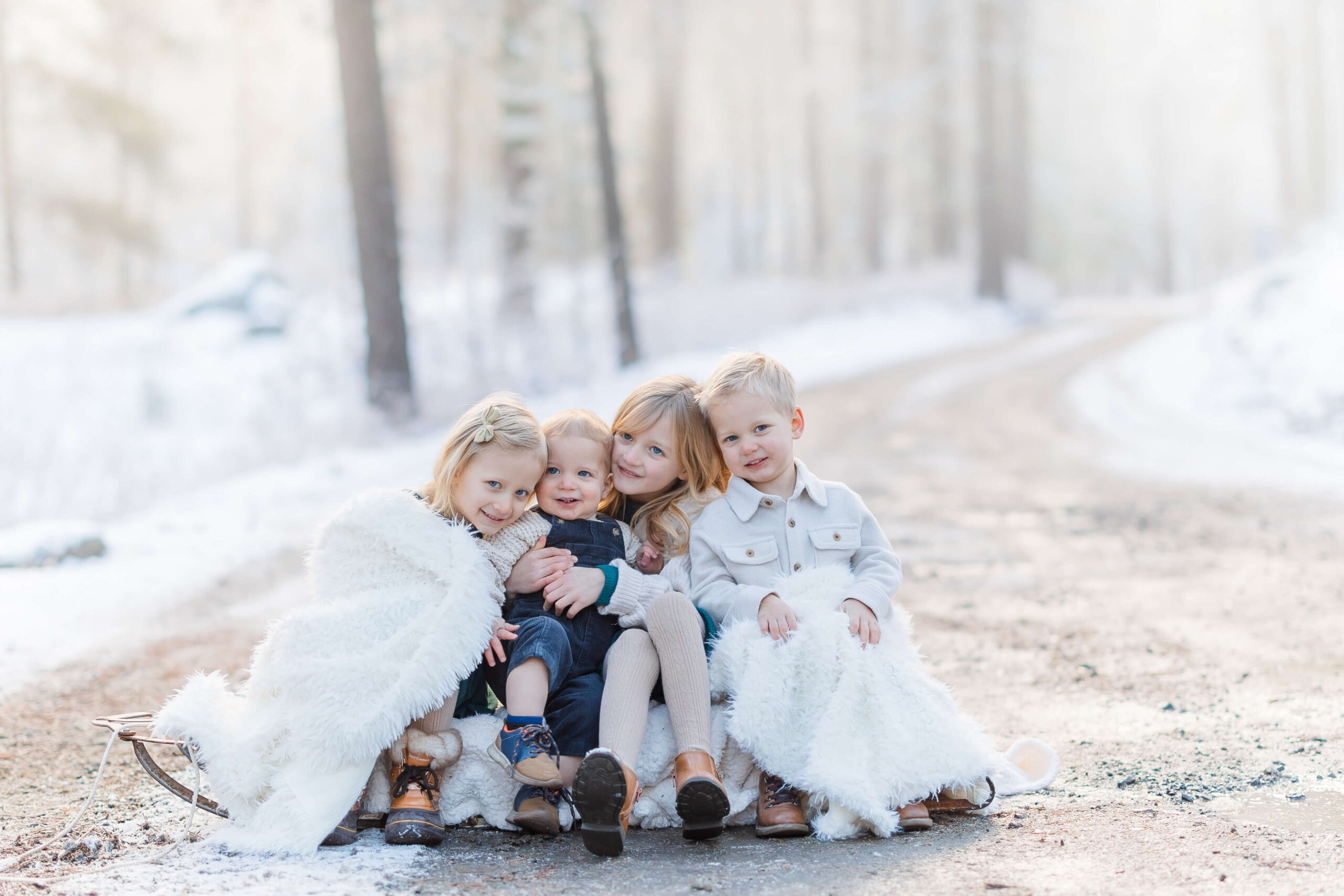 Four young siblings sit on an old sled in a snow covered forest on a gravel road