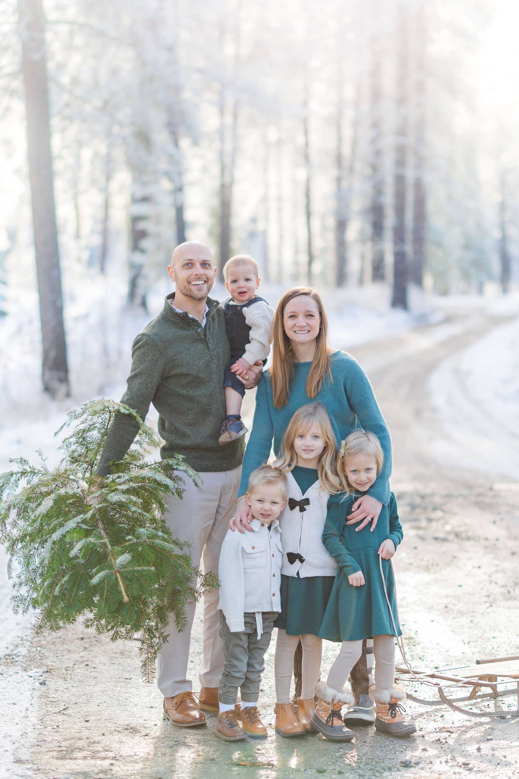 A happy mom and dad stand with a small christmas tree in a forest road with their four children after visiting Leavenworth Reindeer Farm