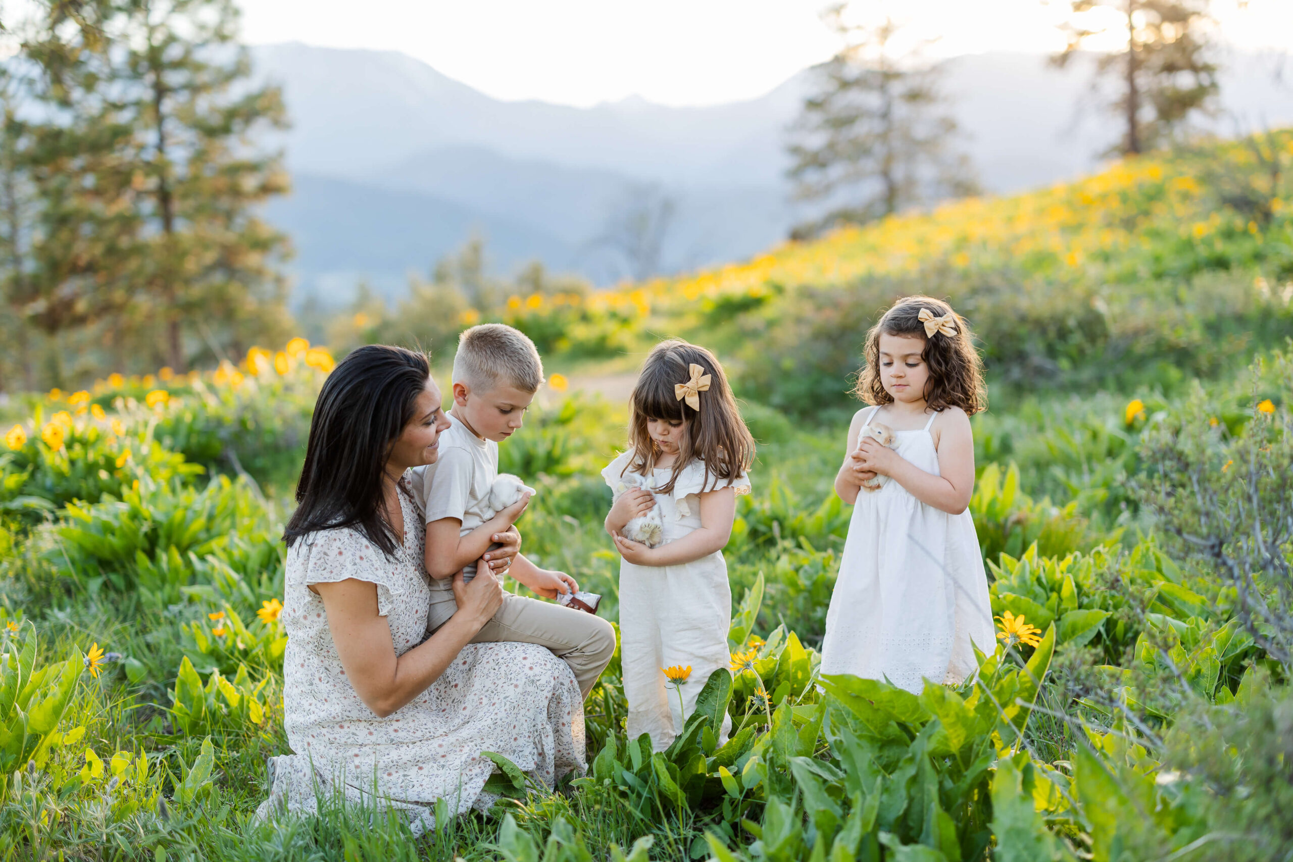 A mom plays with baby bunnies with her three young children in a field of wildflowers in white outfits