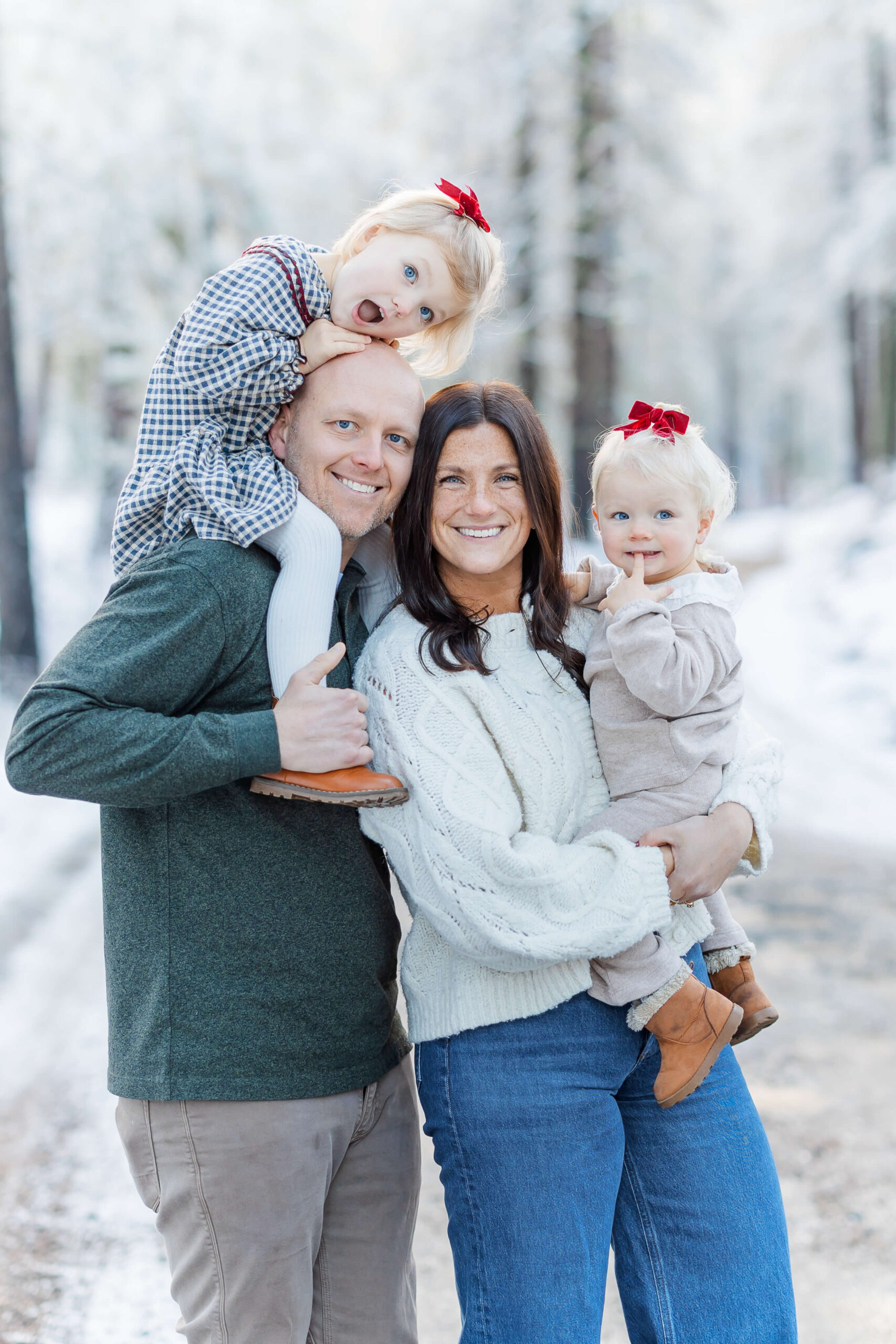 A mom and dad stand in a snow covered forest smiling with their two toddler daughters on shoulders and hip after discovering kid-friendly restaurants in Leavenworth, WA