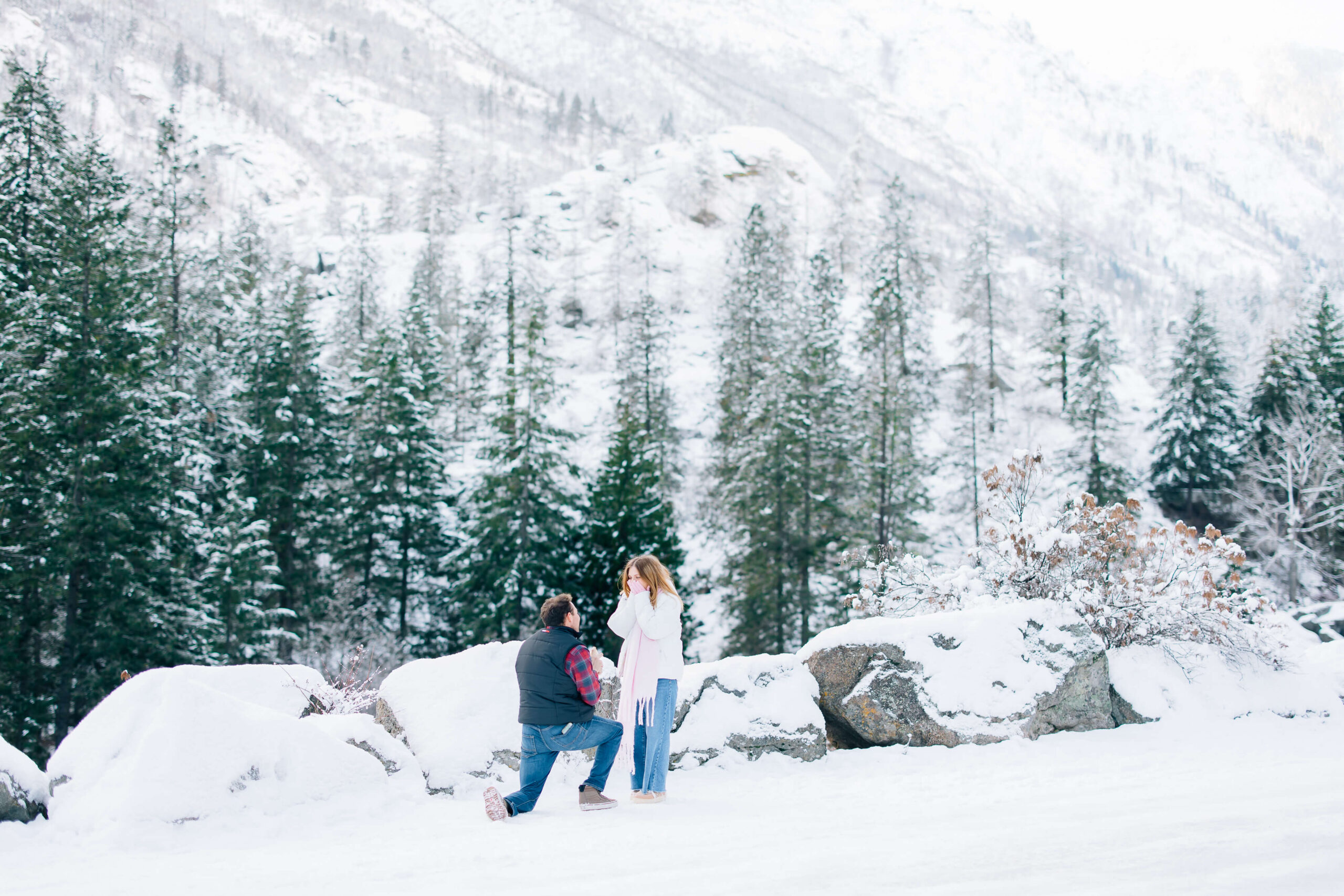 A man in a black vest gets on one knee to propose in the snow covered mountains to his fiancee in white and jeans