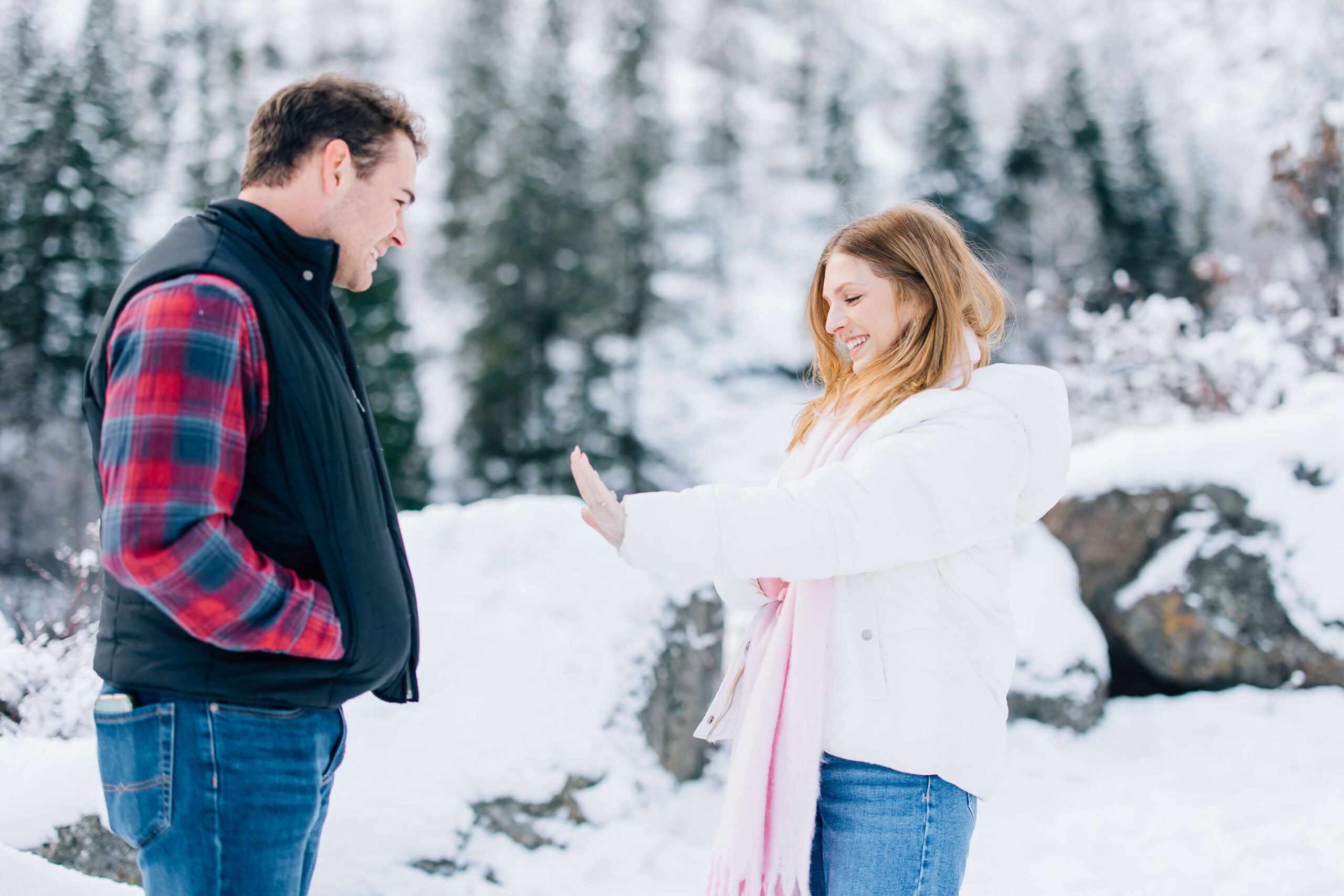 A woman in a white coat and pink scarf admires her new engagement ring as fiancee looks on in snowy mountains after their leavenworth proposal