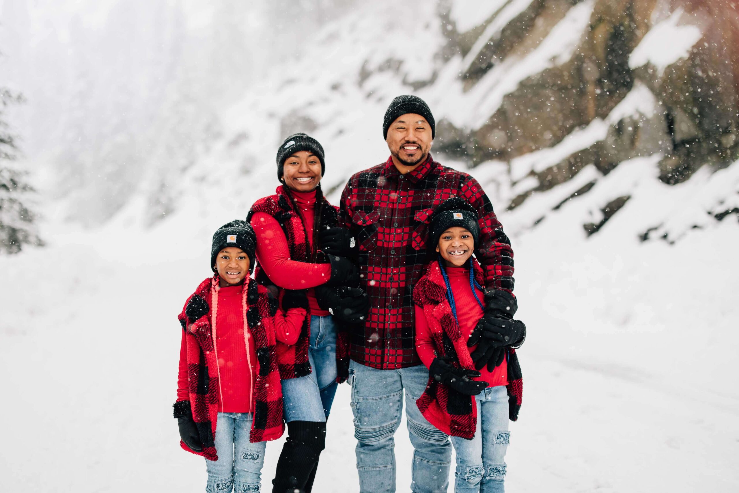 A mom and dad smile while standing int he snow with their young son and daughter in matching red plaid and denim