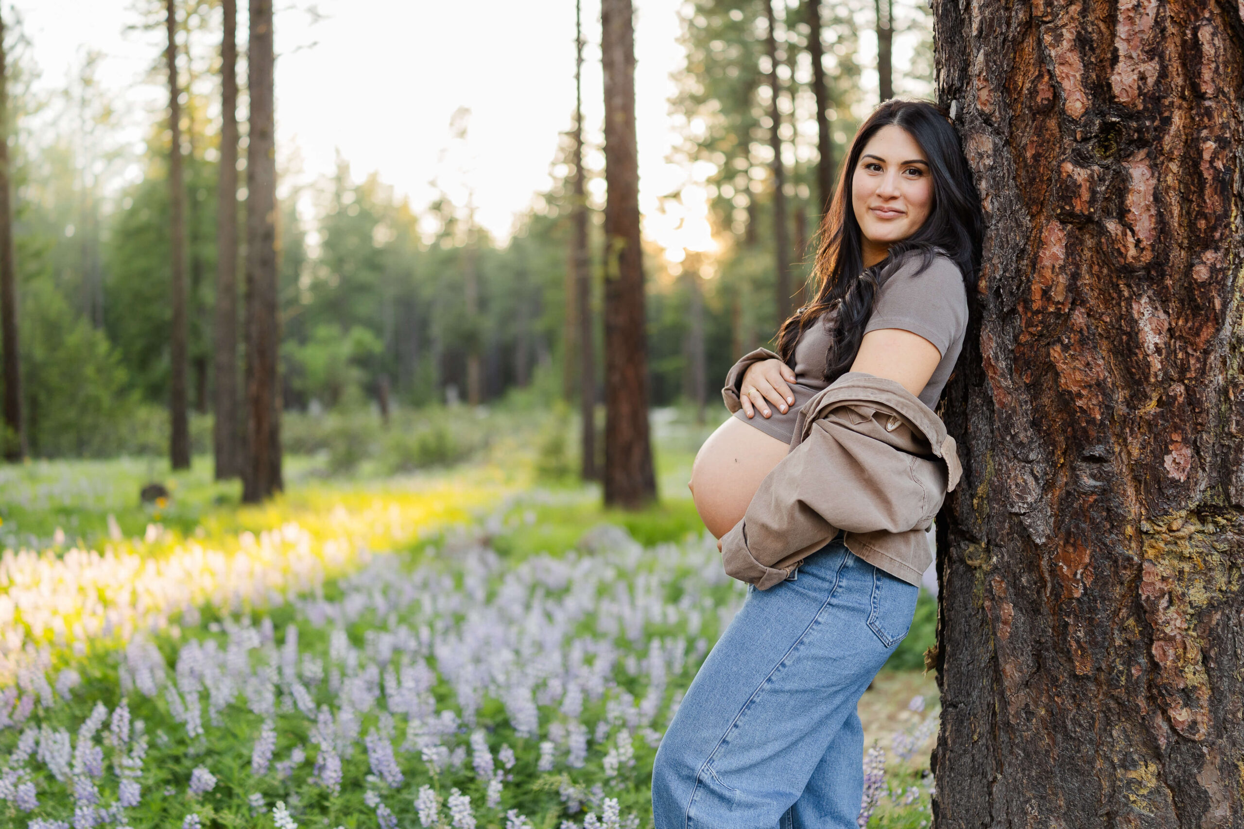 A smiling mother to be in a brown top and jeans leans against a tree with hands on her bump in a forest at sunset