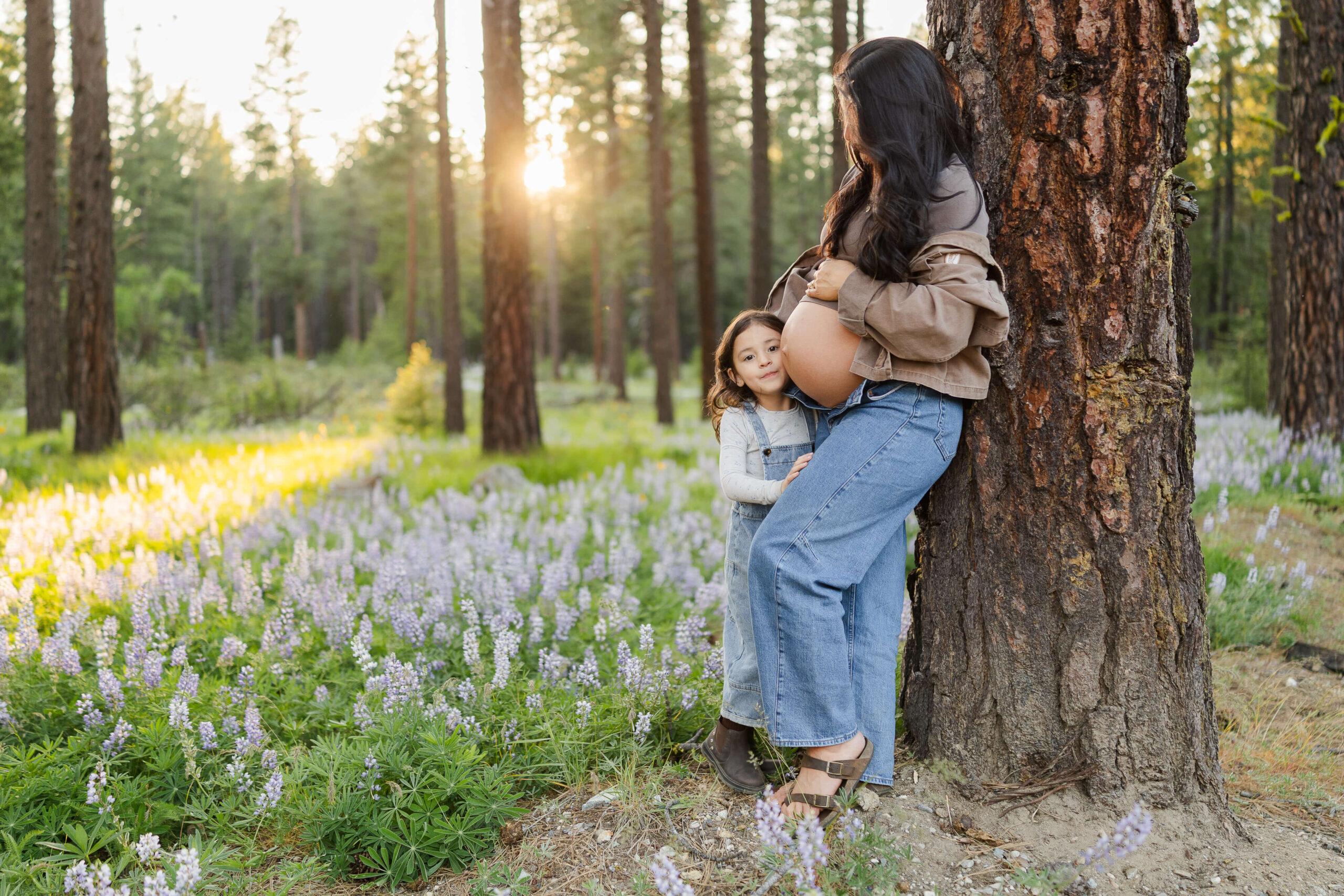 A toddler girl in overalls hugs her pregnant mom leaning against a pine tree in a forest filled with wildflowers after meeting a wenatchee midwife