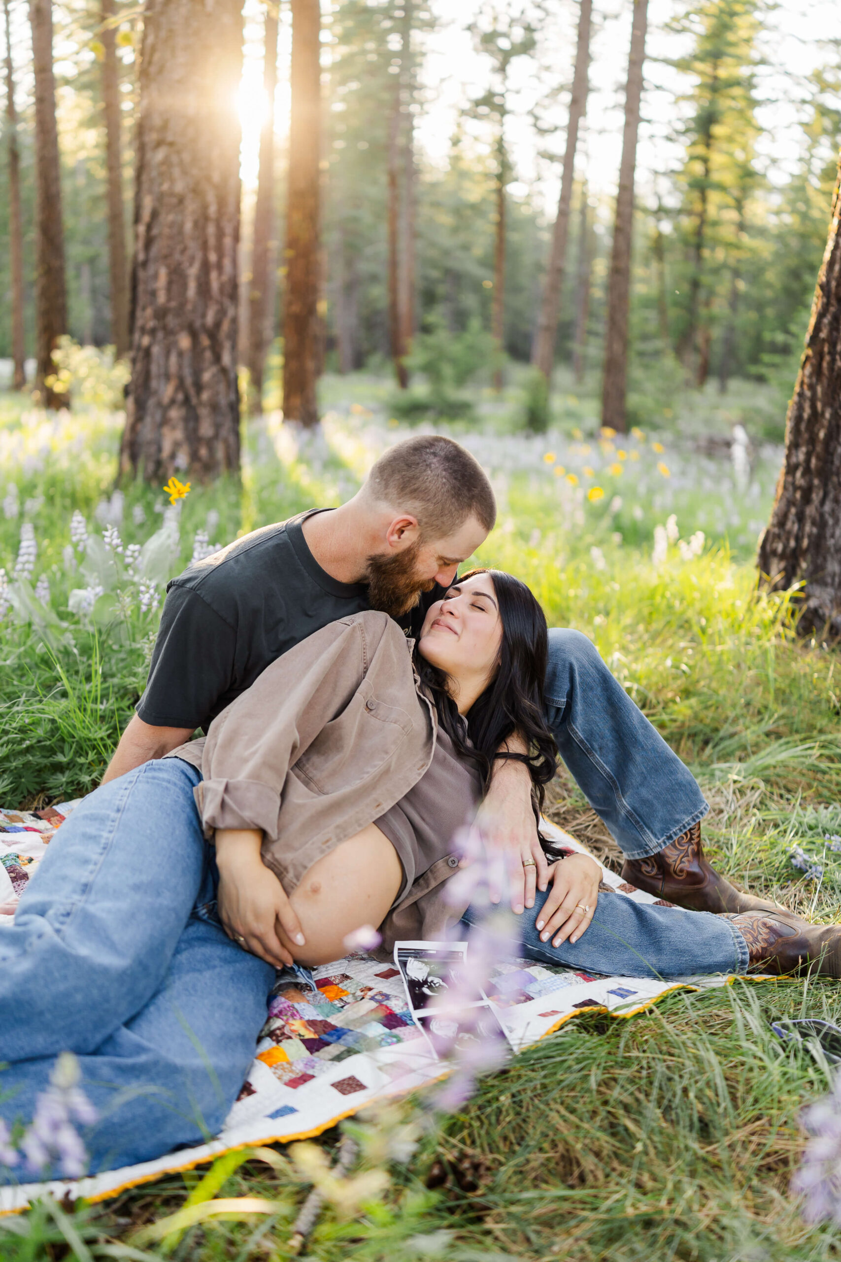 A happy mother to be lays across a picnic blanket in a forest at sunset in the lap of her husband after they found a wenatchee midwife
