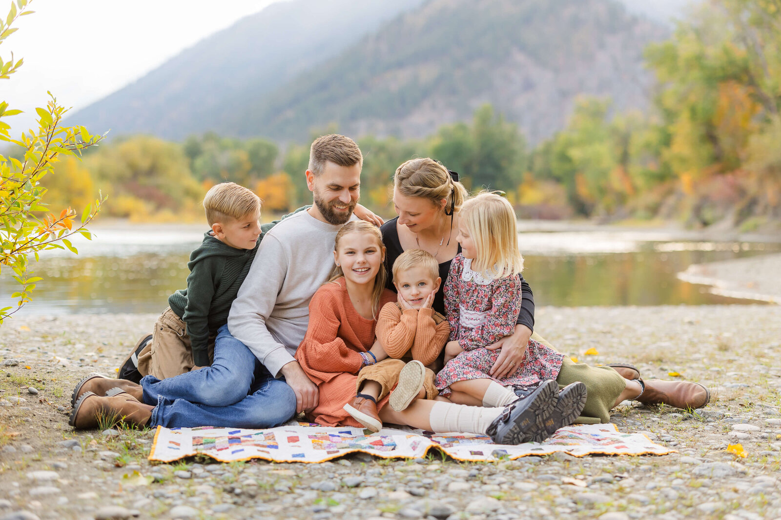 A happy family of six cuddle on a picnic blanket on the river's edge in fall