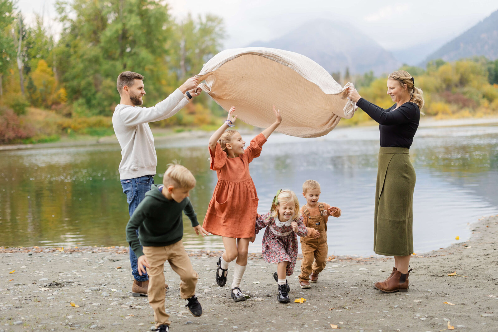 Four young siblings run under a picnic blanket held by mom and dad while playing by a riverbank before visiting the leavenworth autumn leaf festival
